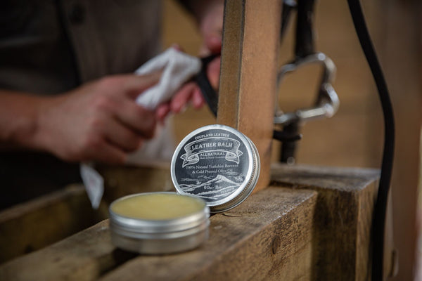 Close-up of Coupland Leather Balm tin beside a vegetable-tanned leather wallet, showing natural beeswax and olive oil ingredients used for conditioning and protection.